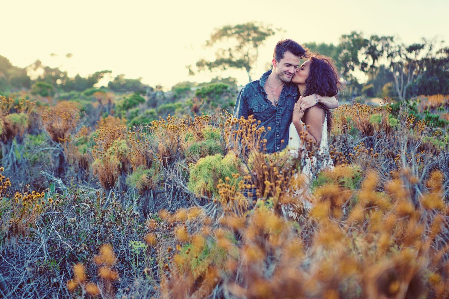 Couple embracing romantically among golden wildflowers in a sunlit field at dusk.