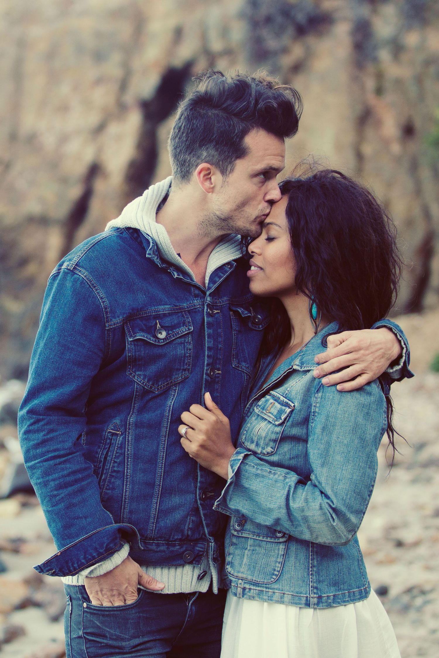 Couple in matching denim jackets sharing a romantic kiss outdoors near rocky cliffs.