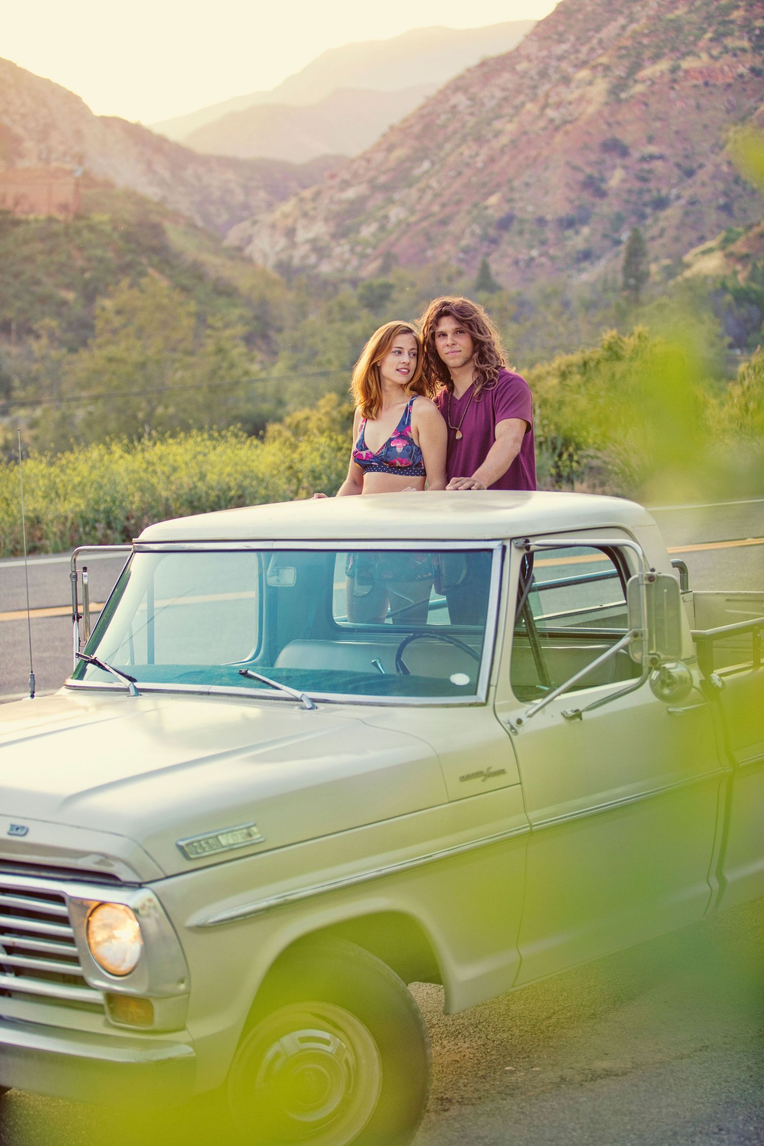 Two people standing through sunroof of vintage white pickup truck with mountain backdrop in golden light.