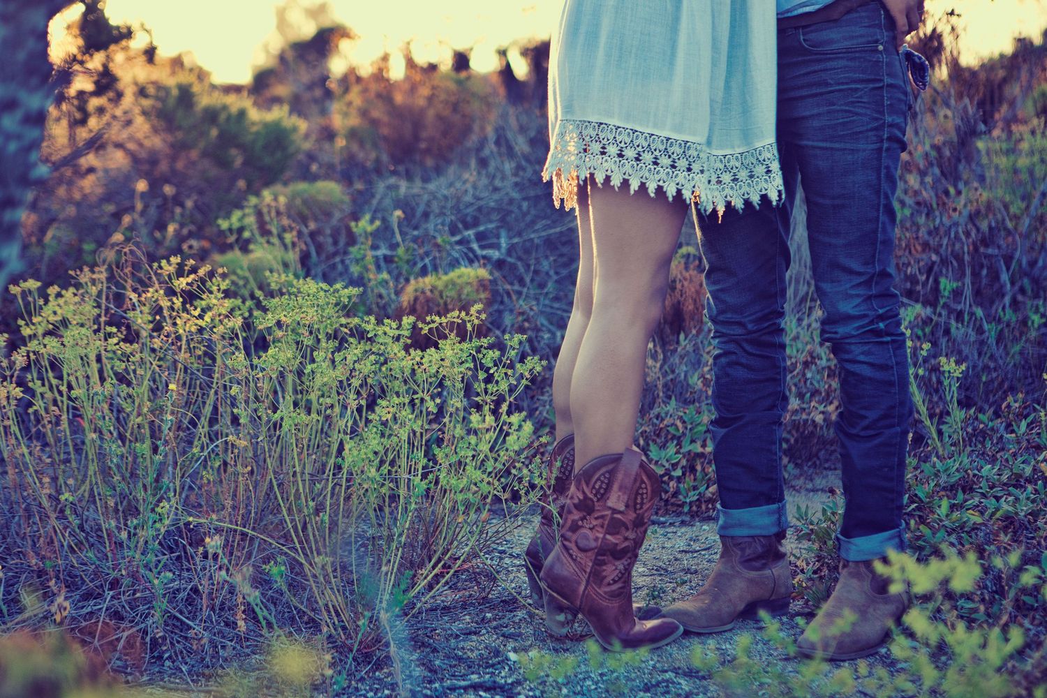 Couple standing close together in a field, wearing jeans and brown boots with a lace dress hem visible.