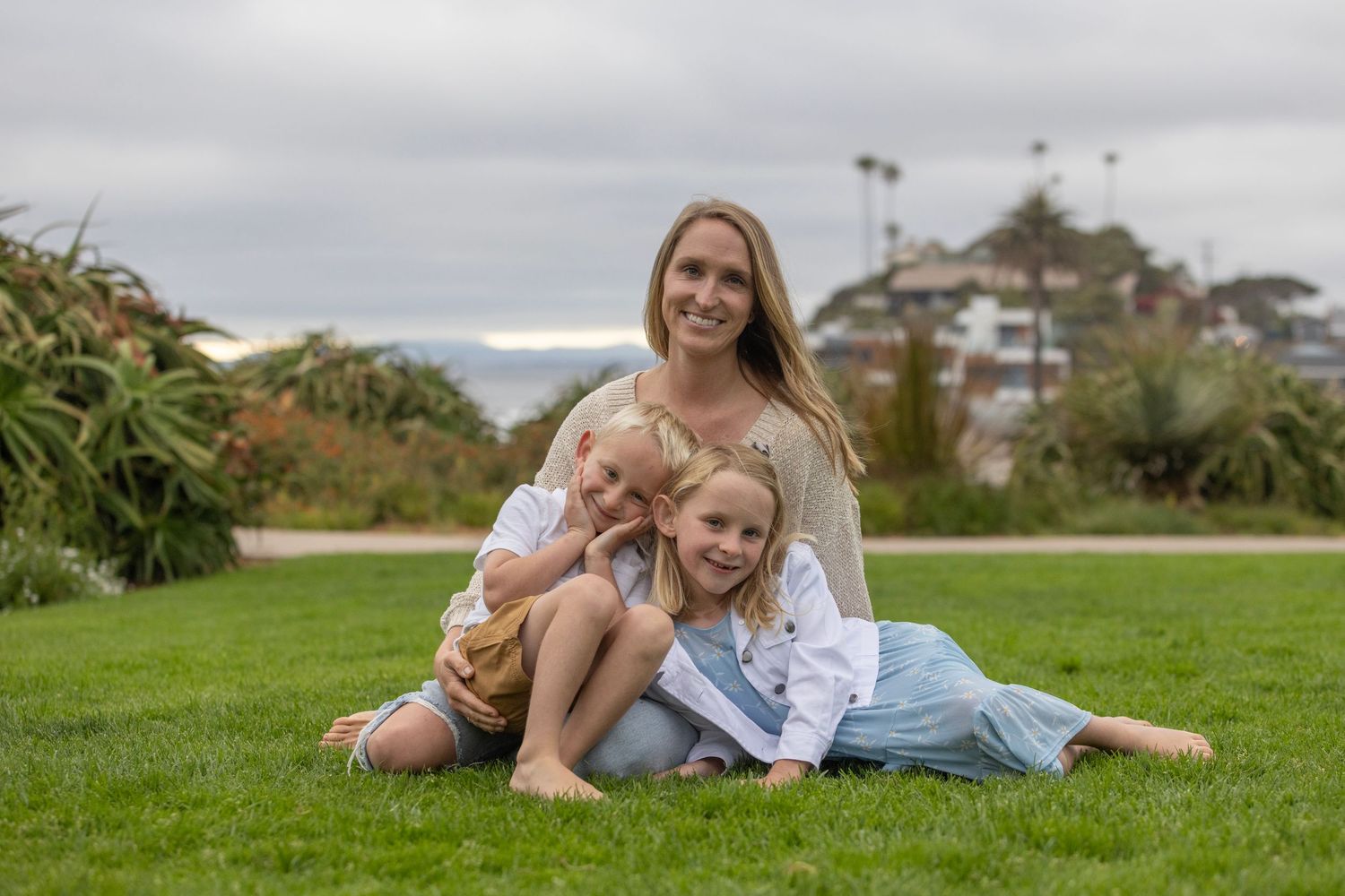 A smiling woman sits on green grass with two young children in a coastal park on a cloudy day.