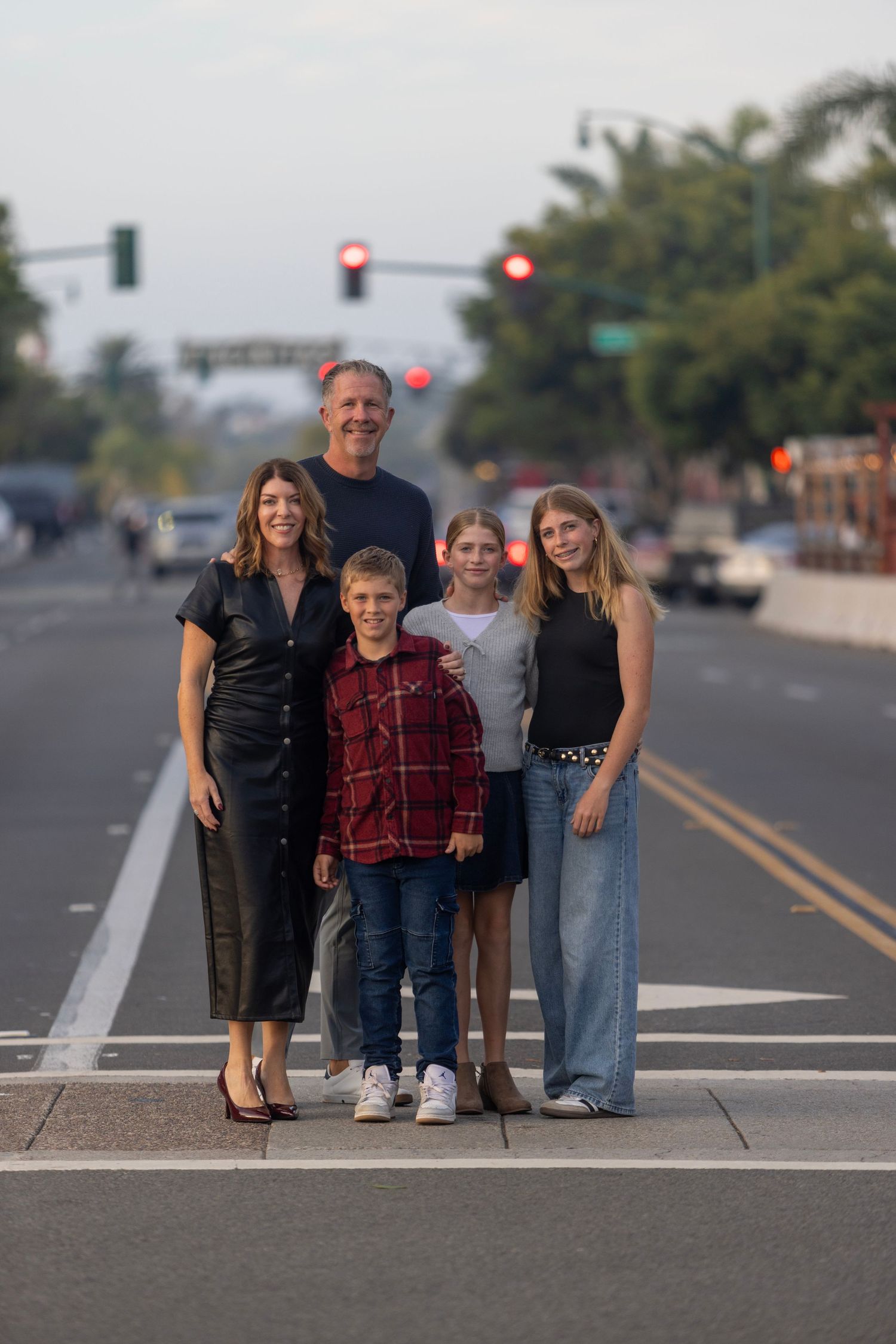 Family of five poses together on a median strip at dusk, with a tree-lined street and red traffic lights behind them.
