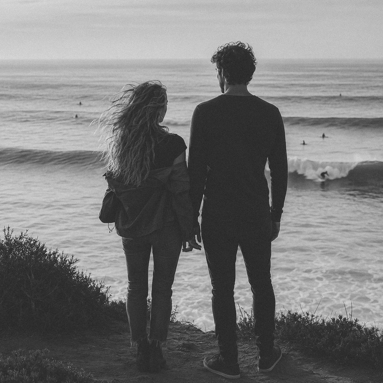 Black and white photo of a couple standing on a coastal cliff overlooking the ocean with surfers in the water.
