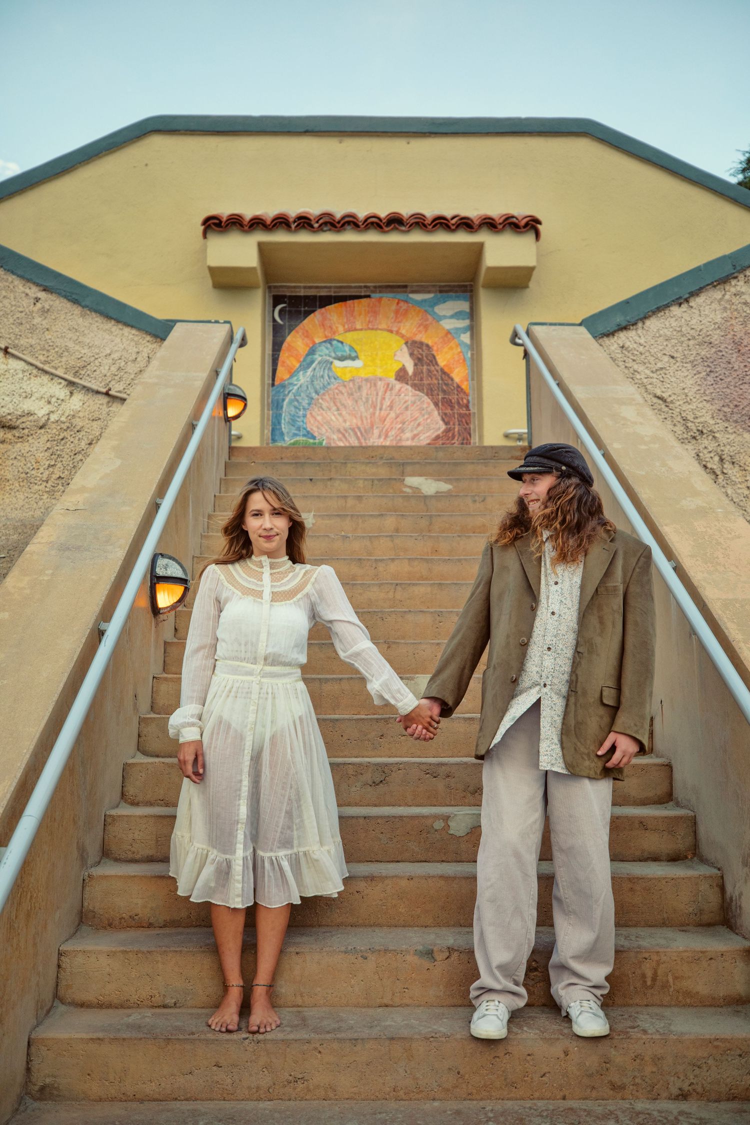 Couple holding hands on stone steps outside a colorful mosaic building, woman in white dress, man in casual earth tones.
