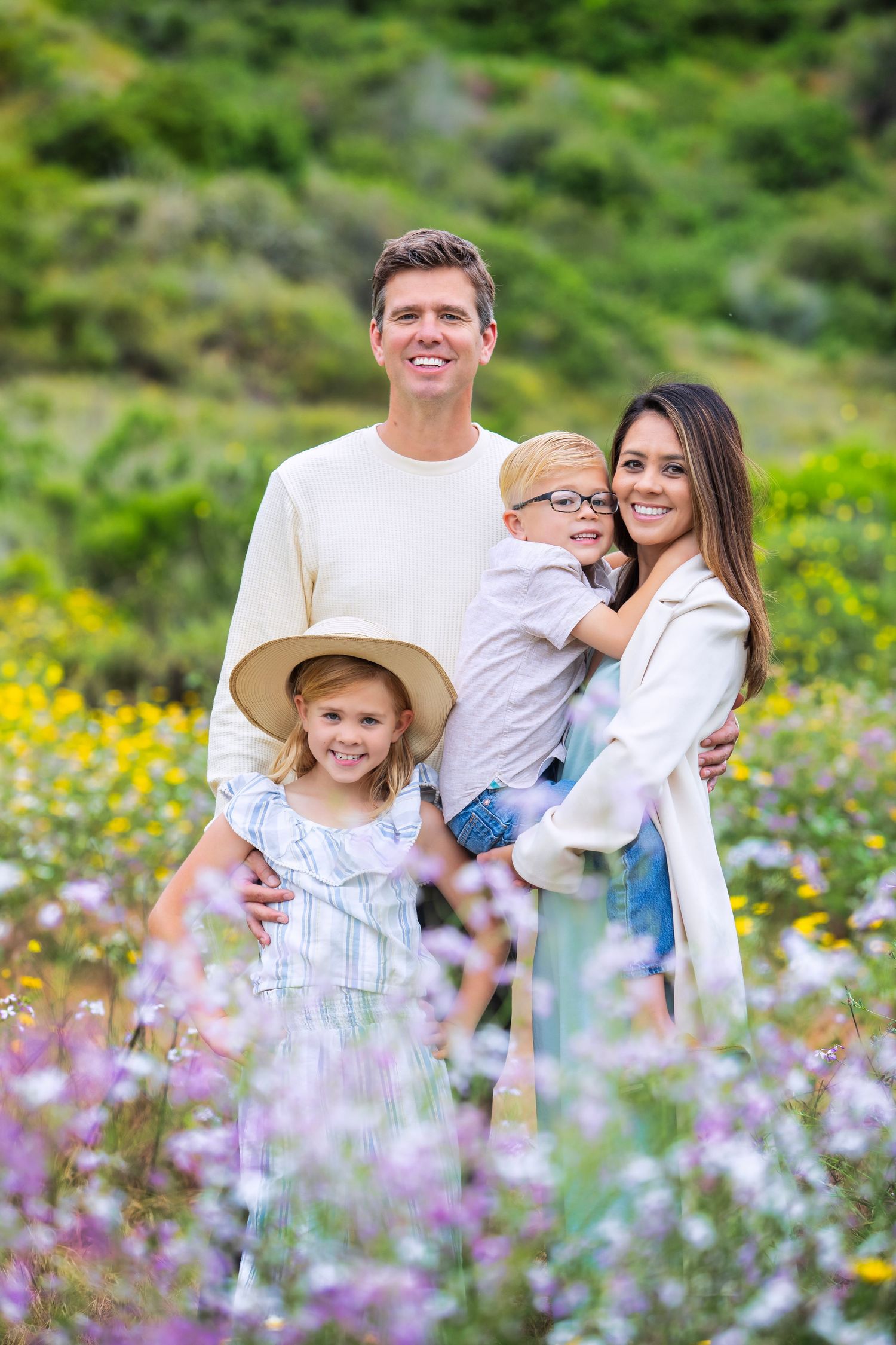 Happy family of four posing in a colorful wildflower meadow, parents smiling with two young children outdoors.