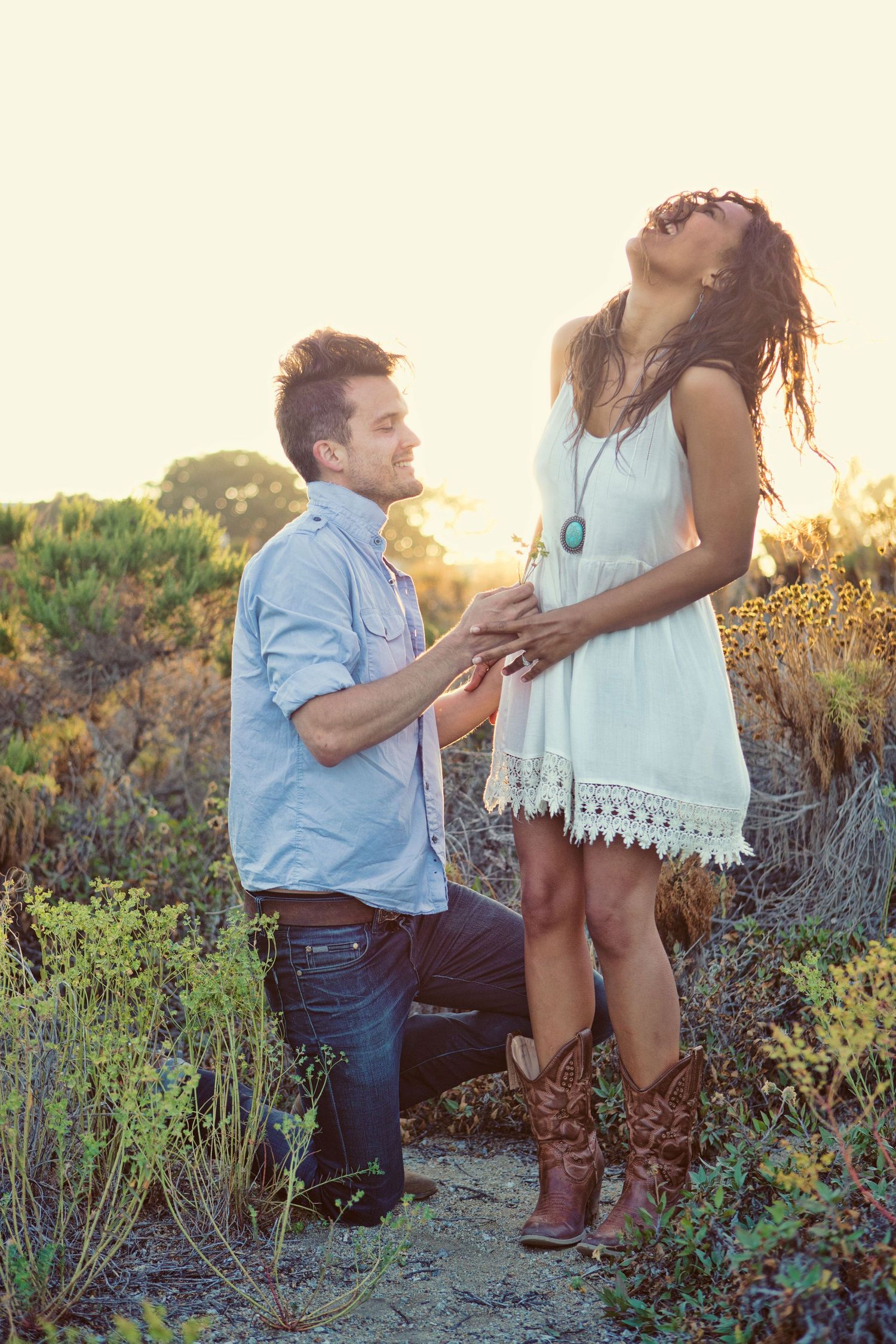 Man proposes on one knee to woman in white dress and cowboy boots outdoors at golden sunset.