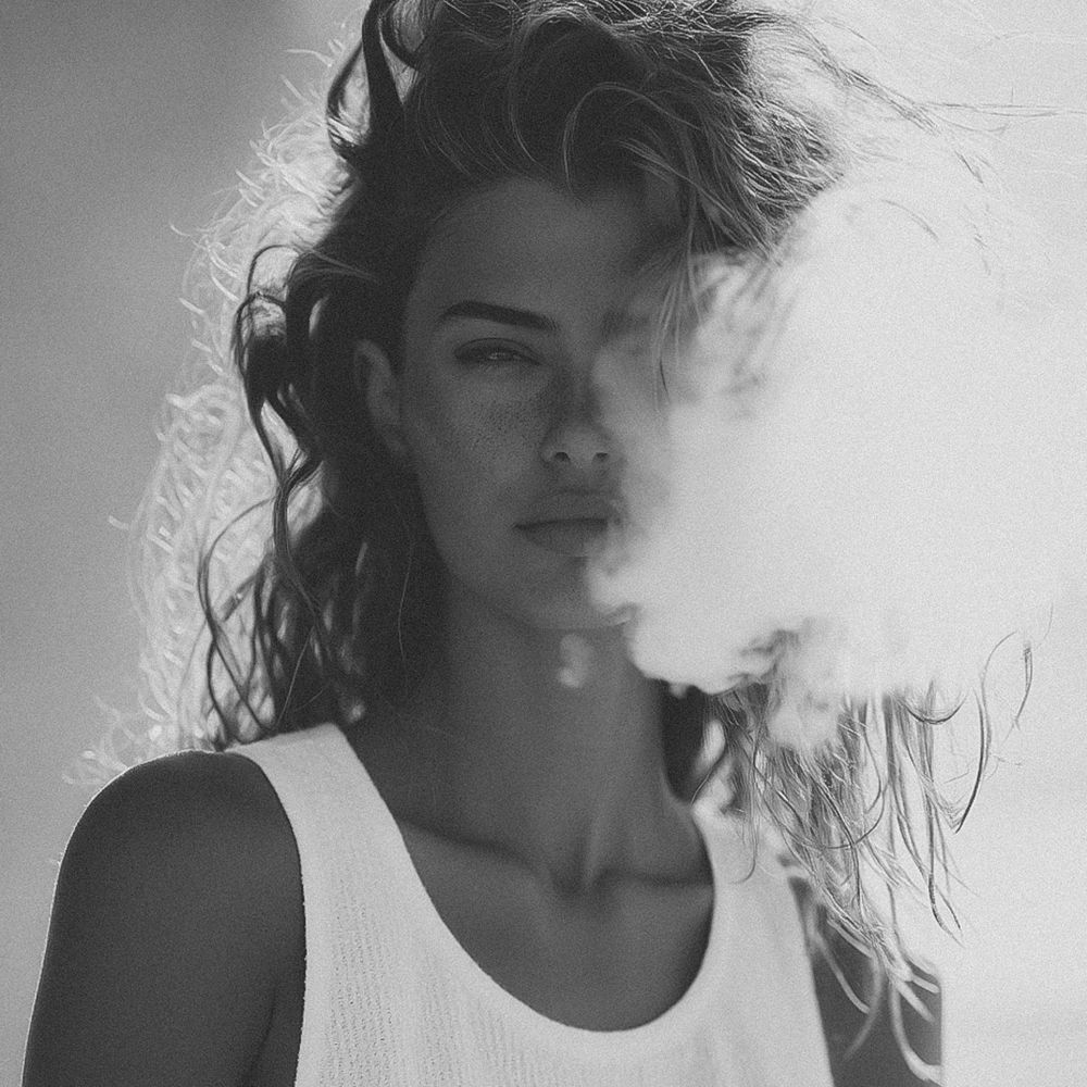 Black and white photo of a young woman with tousled hair exhaling smoke, wearing a white tank top.