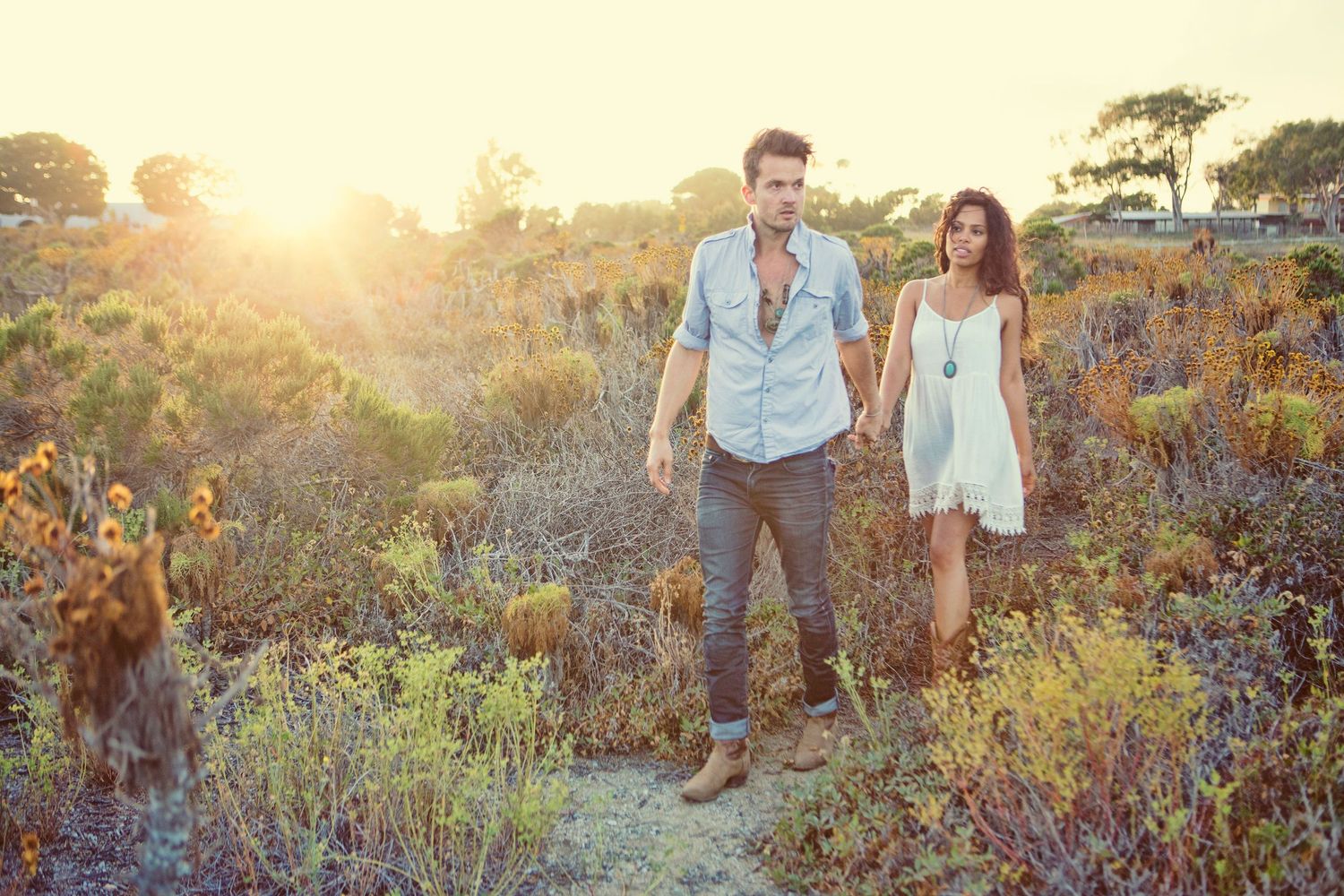 Couple holding hands walking through golden sunlit shrubland, man in denim shirt and woman in white dress.