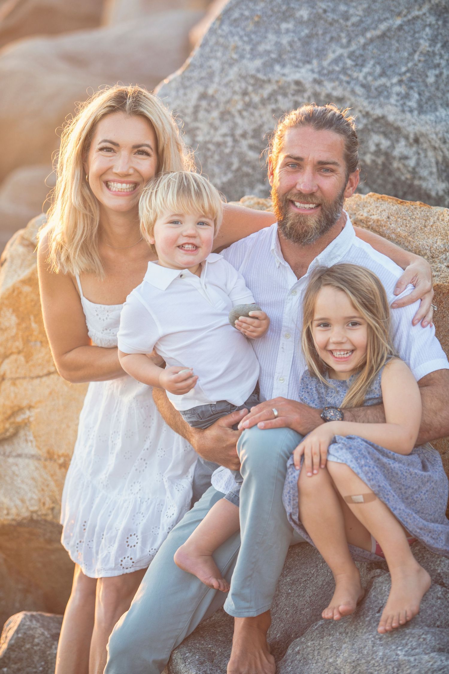 Happy family of four posing on rocks at the beach during golden hour, parents smiling with two young children.