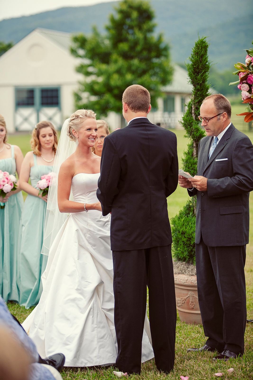 Outdoor wedding ceremony with bride in white gown, groom in black suit, officiant reading vows, bridesmaids in mint dresses.
