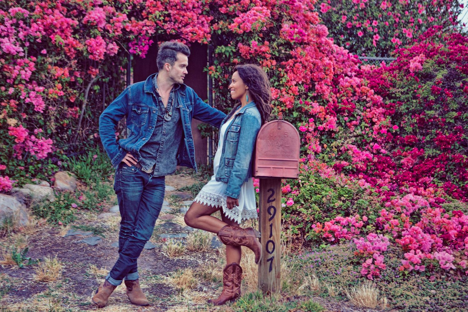 Couple standing by vintage mailbox surrounded by vibrant pink bougainvillea flowers in denim outfits.
