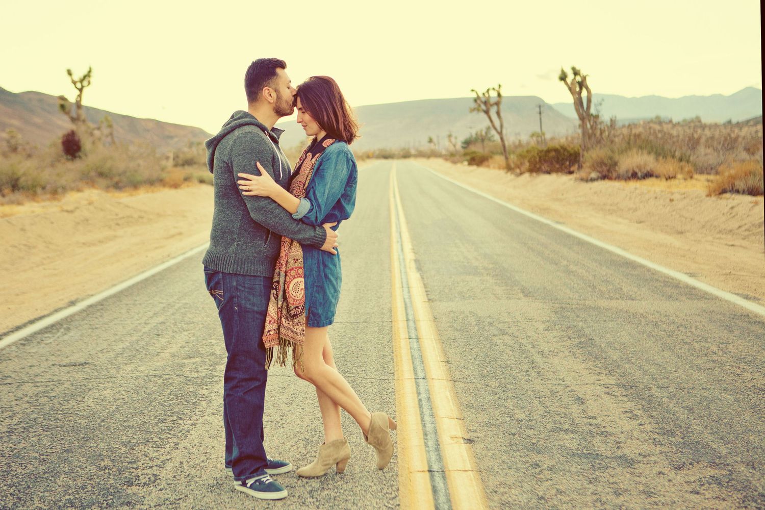 Couple sharing a romantic kiss on an empty desert highway with mountains in the background.
