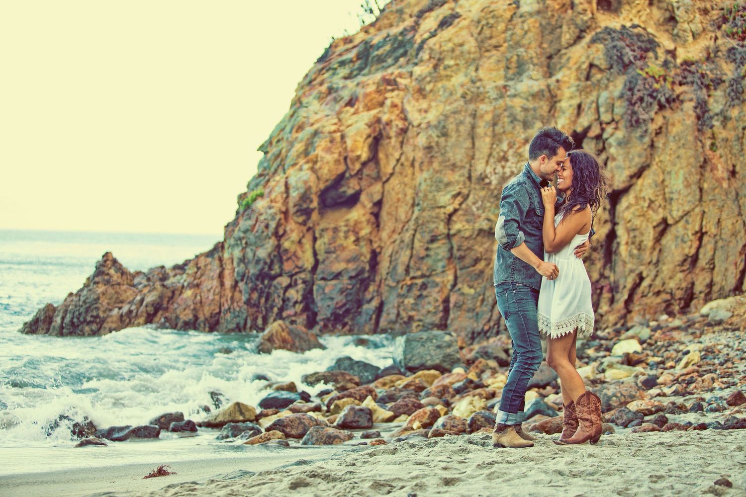 Romantic couple embracing on a rocky beach shore with ocean waves and rugged cliffs in the background.