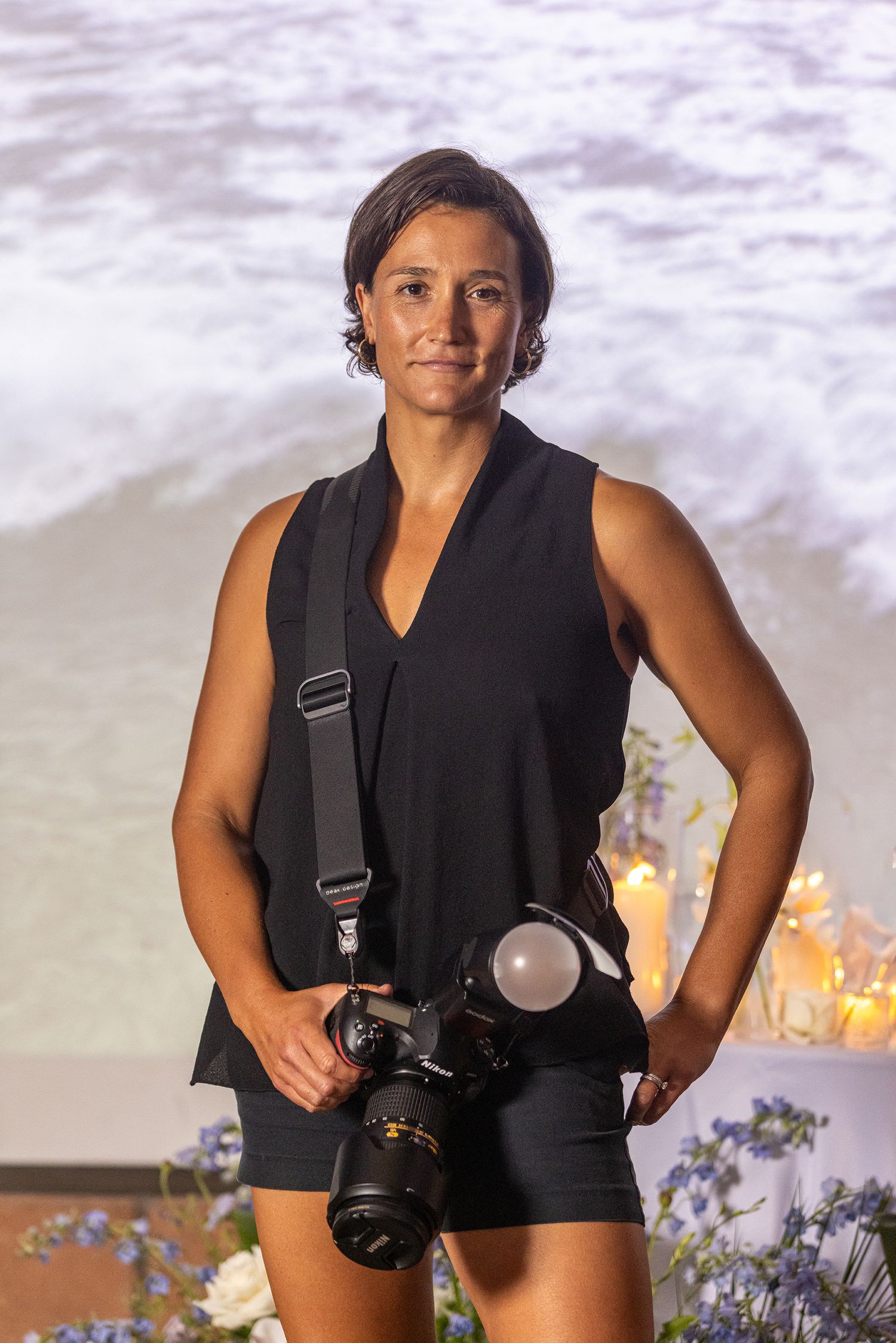 Female photographer in black outfit holding a DSLR camera with flash, standing before a scenic ocean backdrop.