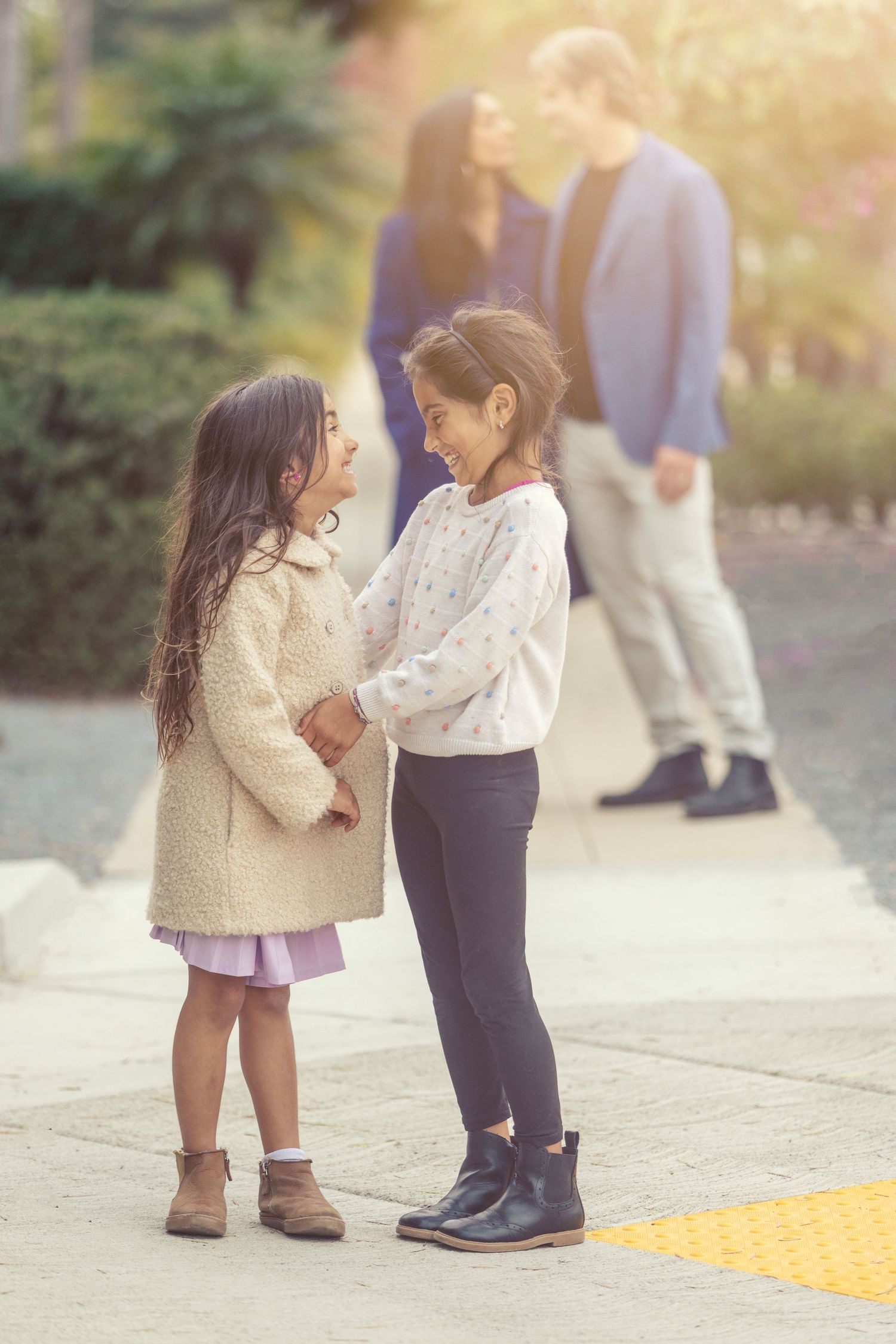Two young girls holding hands and smiling at each other outdoors, with parents standing in the background.