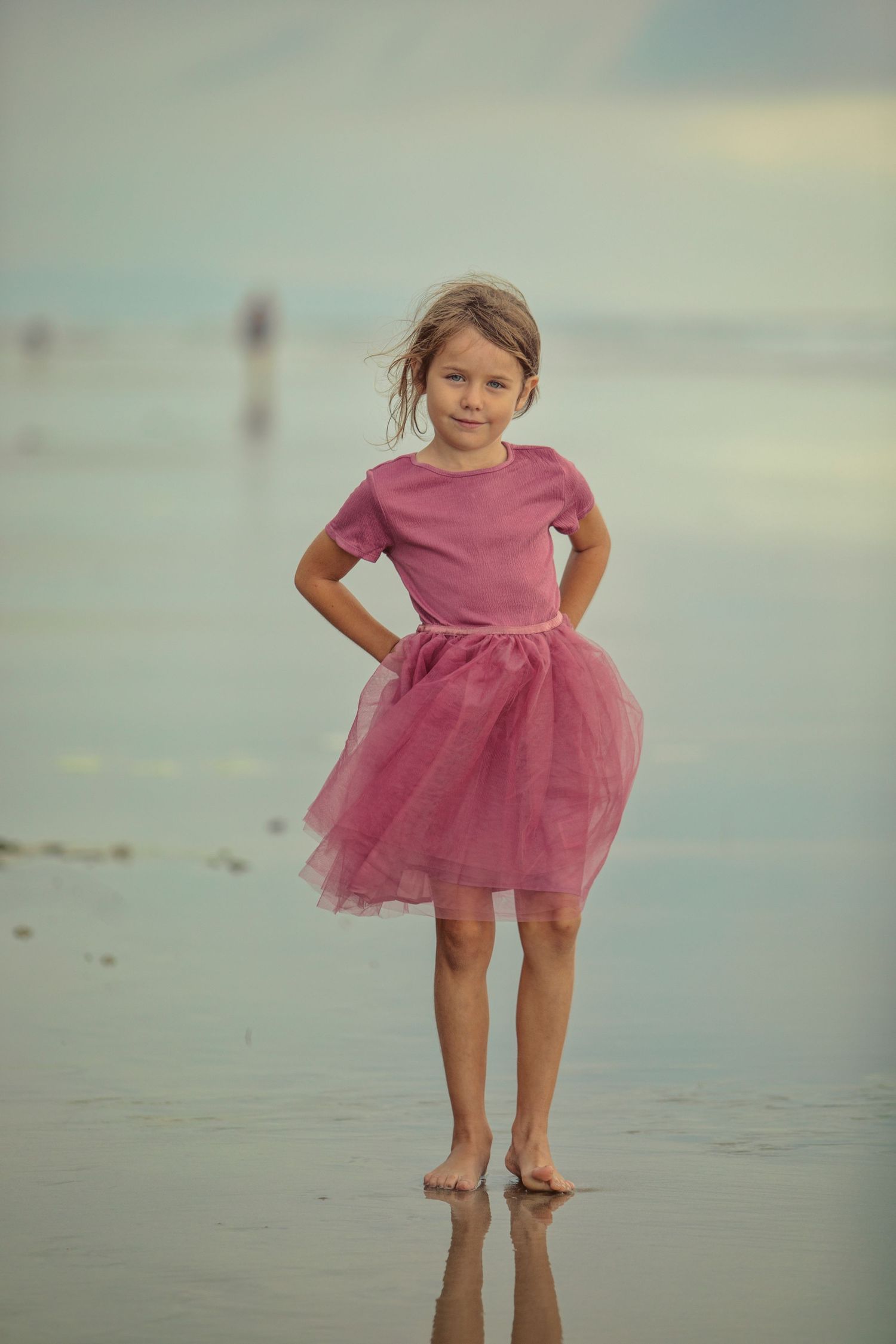 Young girl in pink tutu skirt and top standing barefoot on a reflective beach shoreline.