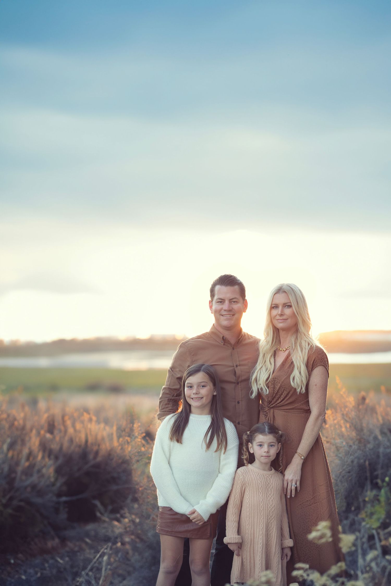 Family of four posing outdoors at golden hour in a field near water, wearing earth tones.