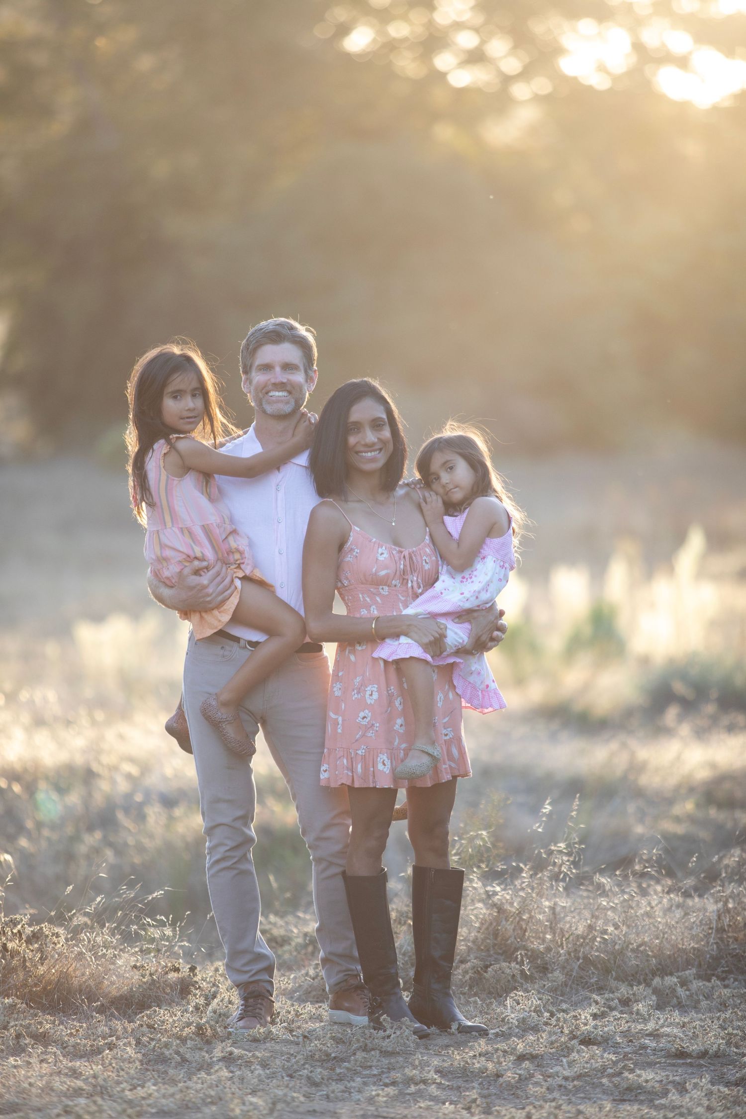 Family of four posing outdoors in golden sunset light, parents holding two young girls in matching pink outfits.