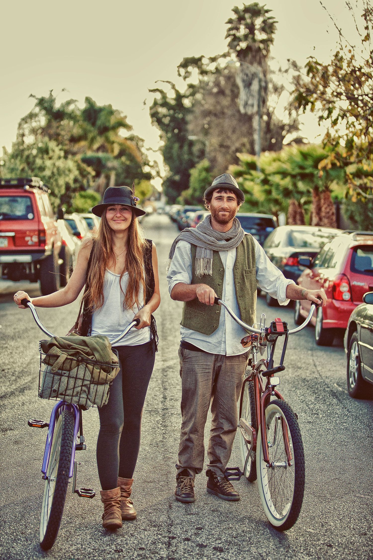 A young couple standing with bicycles on a tree-lined street, wearing casual bohemian outfits and hats.