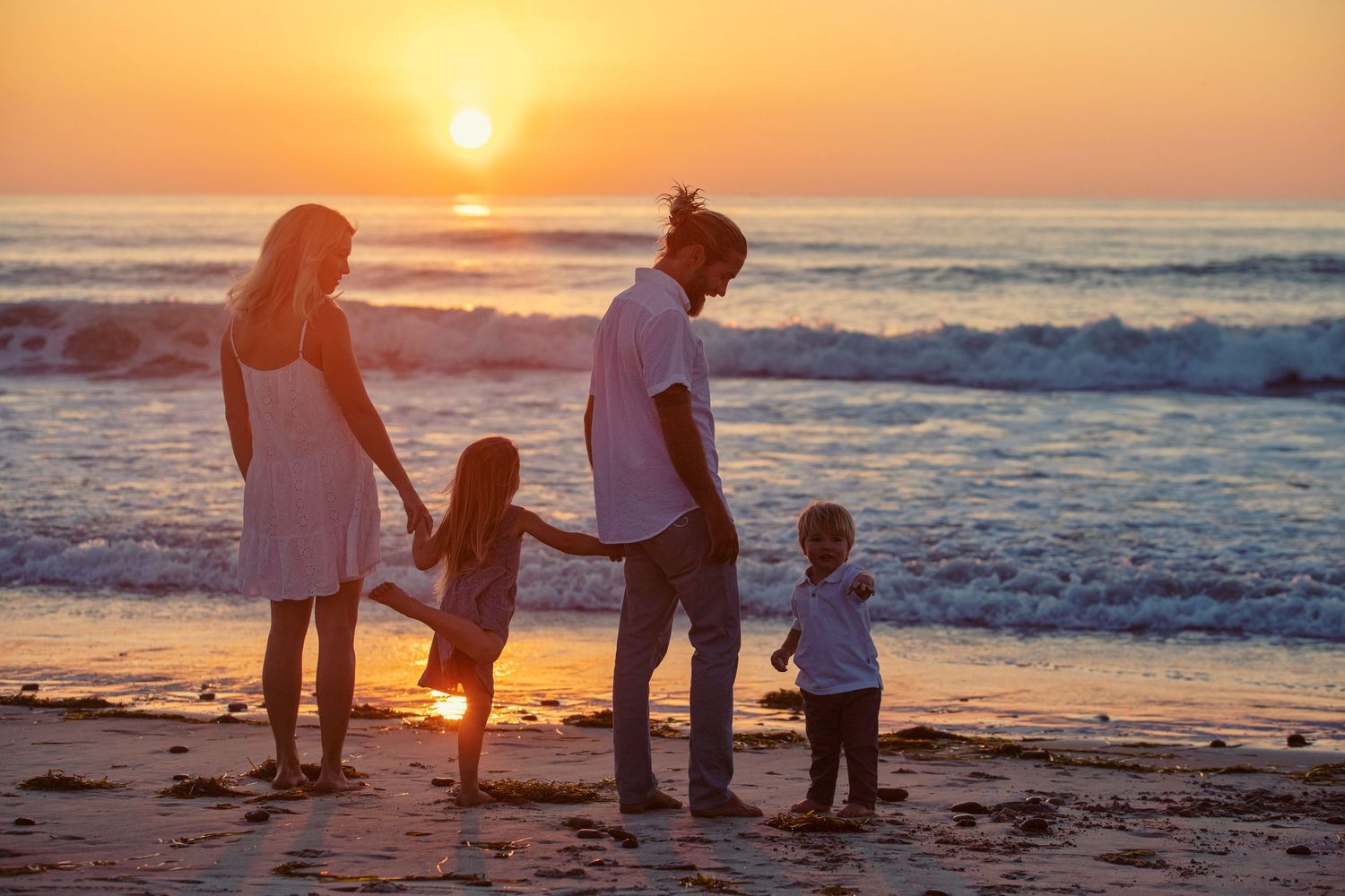 Family of four holding hands on beach at golden sunset, waves in background.
