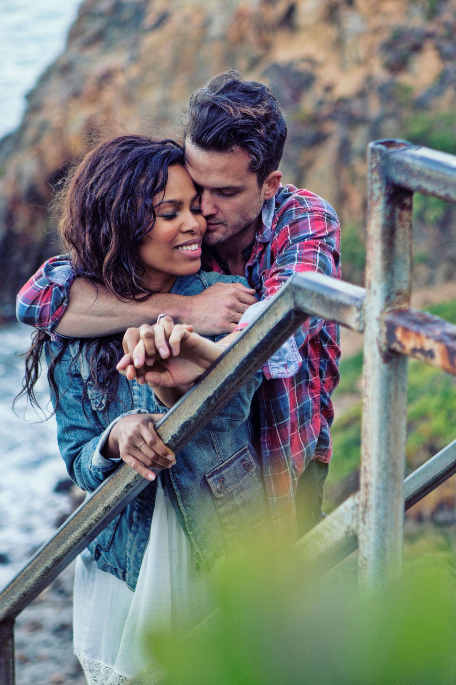 Romantic couple embracing on coastal stairs, woman in denim jacket, man in plaid shirt, rocky ocean backdrop.