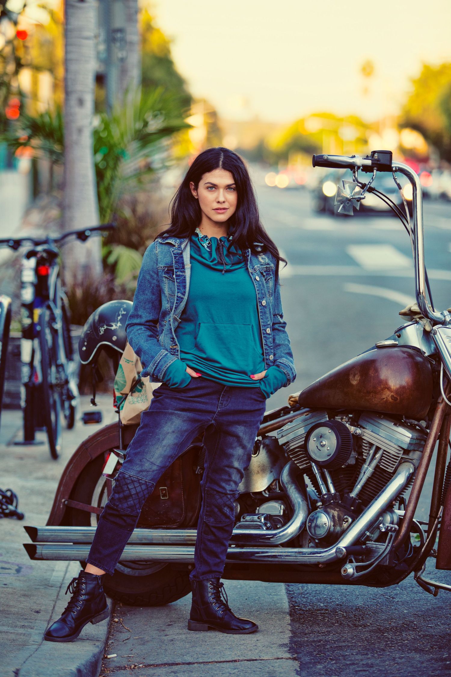 A young woman in a denim jacket and teal hoodie leans against a vintage motorcycle on an urban street.