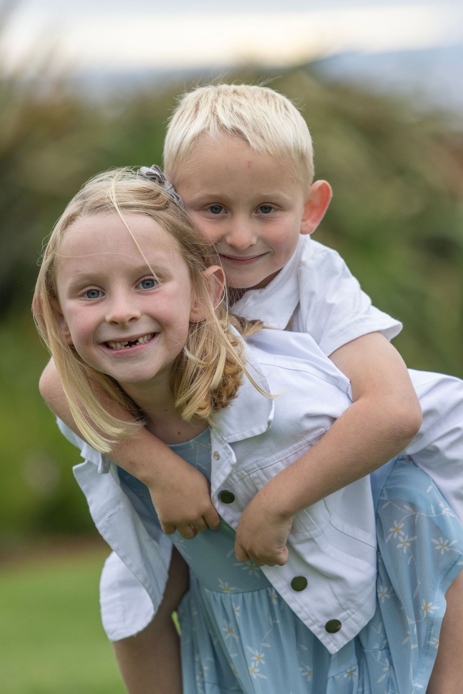 Two blonde siblings smiling outdoors, girl giving boy a piggyback ride in a garden setting.