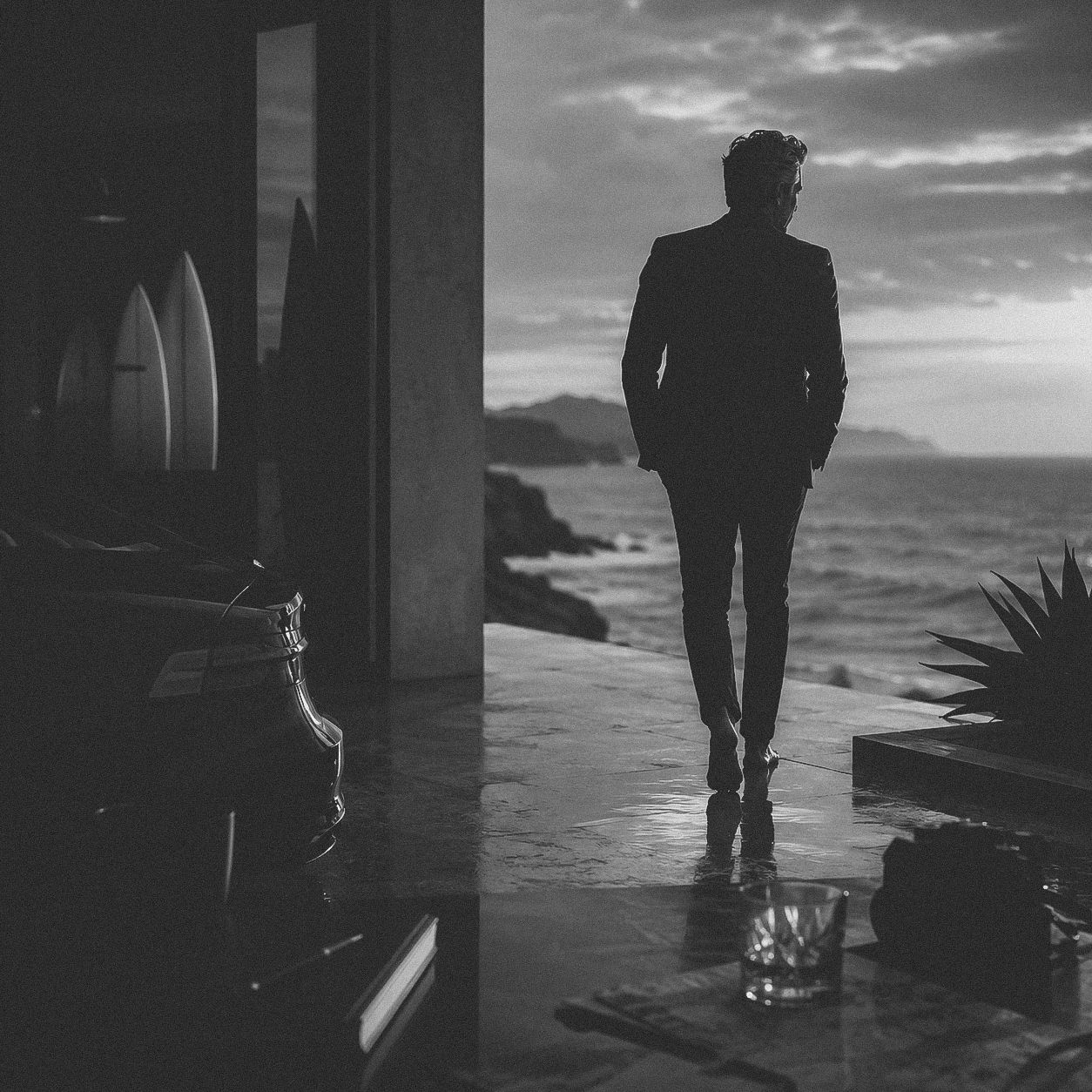 Silhouette of a man in a suit standing on a terrace overlooking a dramatic ocean sunset in black and white.