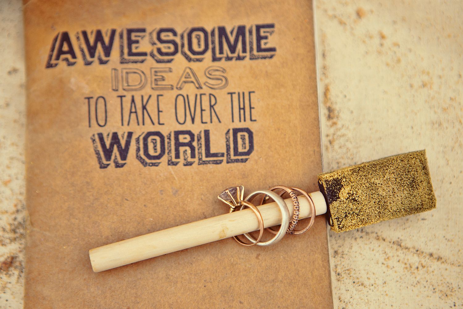 A rustic journal labeled Awesome Ideas to Take Over the World beside a decorative wand with rings on a sandy surface.