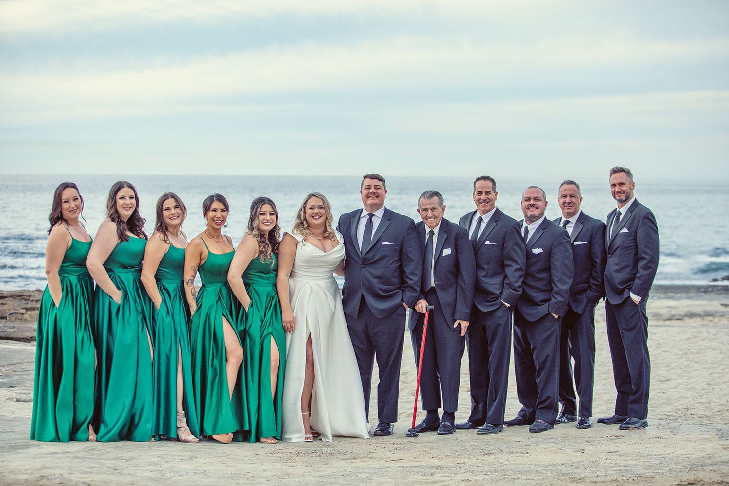 Wedding party poses on beach, bridesmaids in emerald green gowns, groomsmen in navy suits, bride and groom at center.