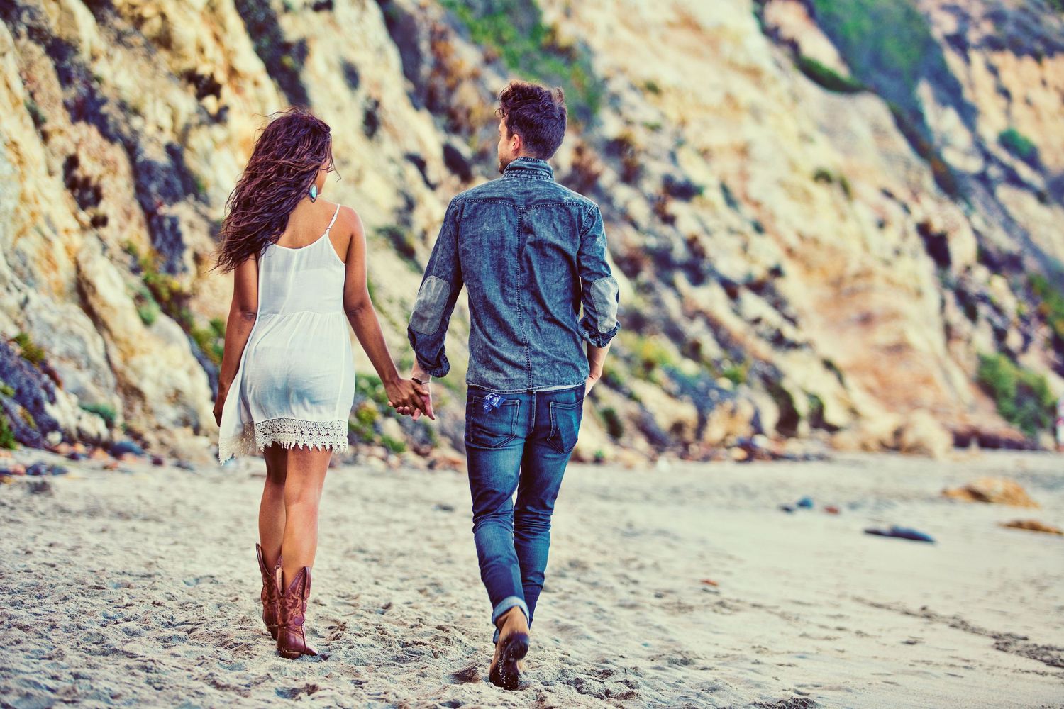 Couple holding hands walking on a sandy beach with rocky cliffs, woman in white dress and man in denim jacket.