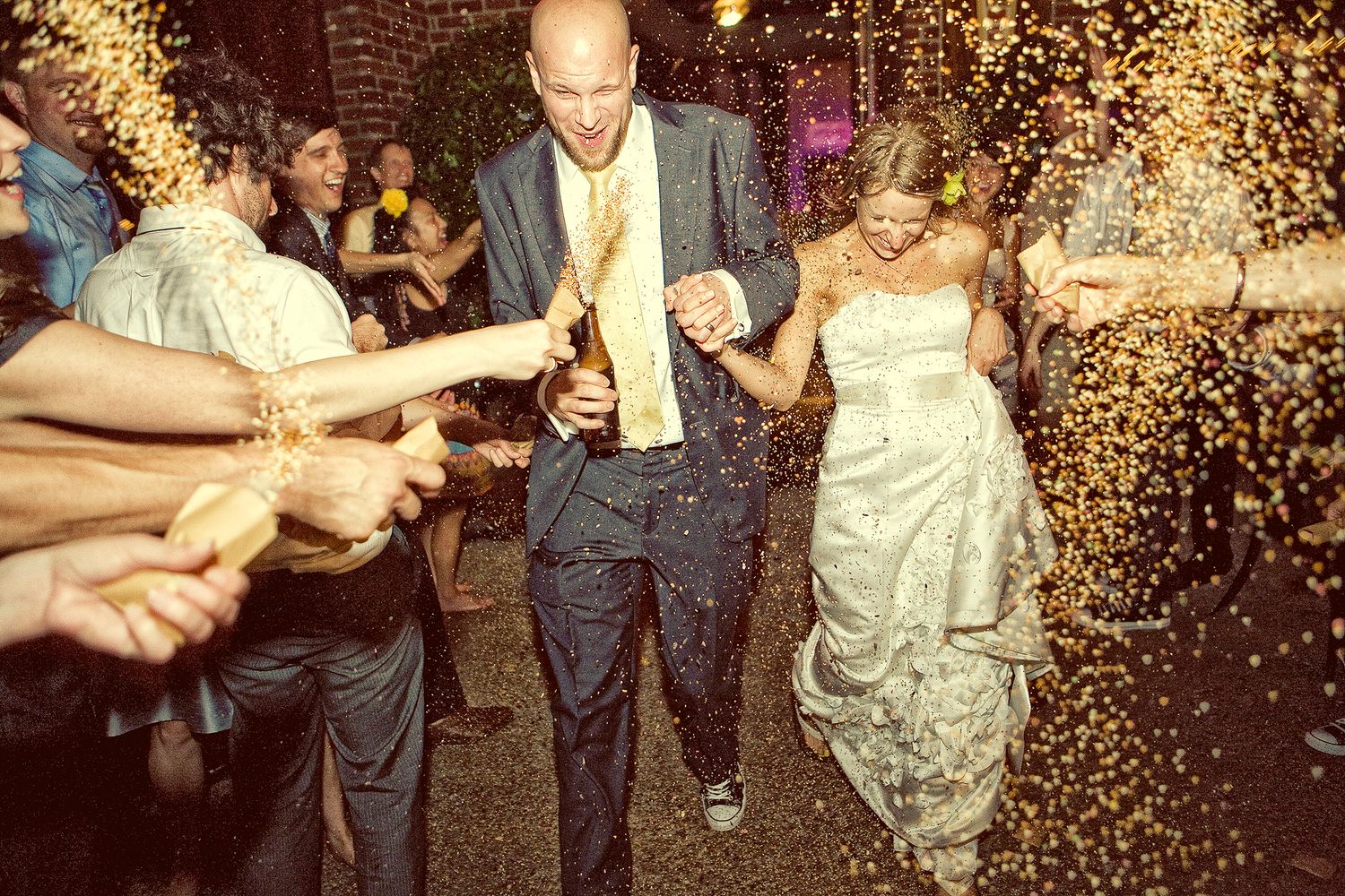 Joyful newlyweds walk through a golden confetti shower during their wedding reception celebration at night.