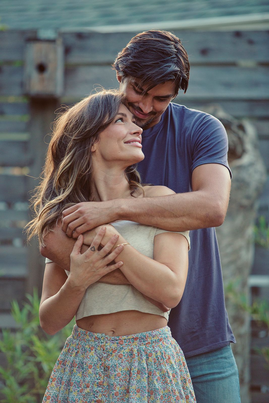 A smiling couple embracing outdoors, man hugging woman from behind near a rustic wooden fence.
