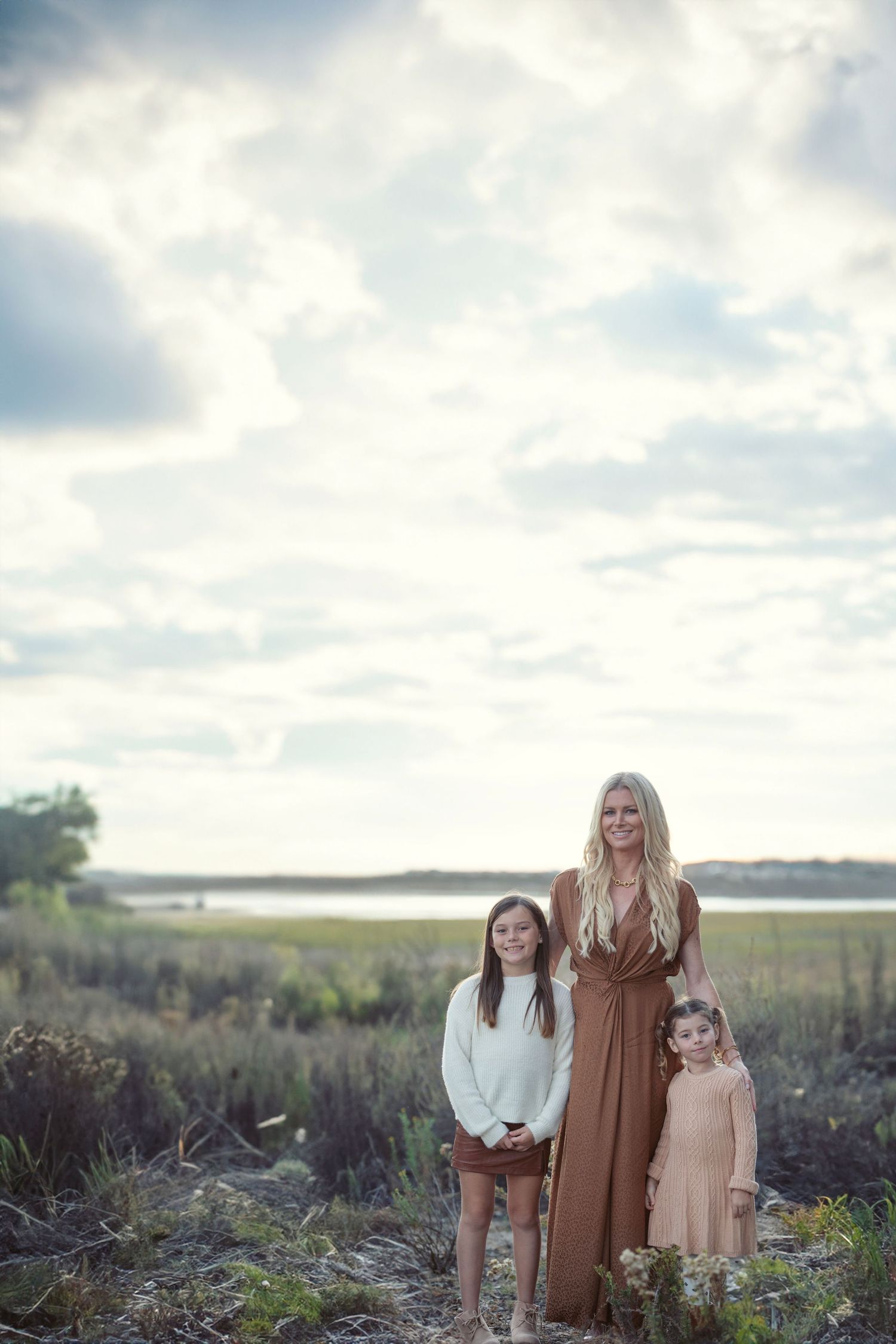 A blonde woman in a brown dress poses outdoors with two young girls in a field near a lake under cloudy skies.