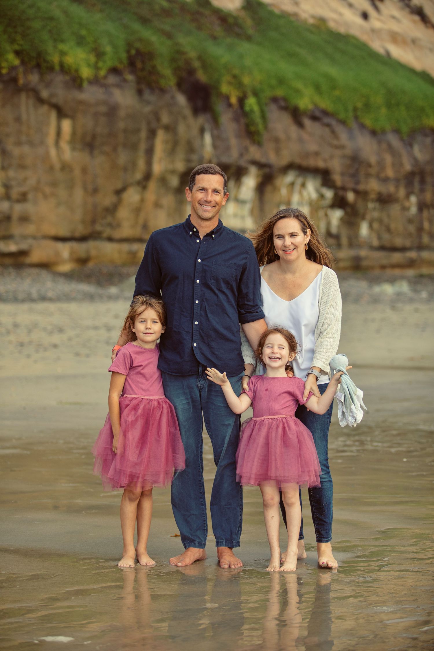 Family of four poses on a sandy beach, two girls in pink tutus with parents, rocky cliffs in background.