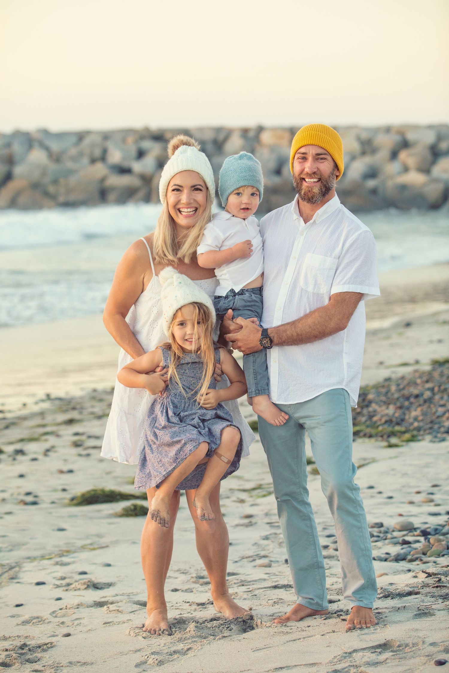 Happy family of four posing on a sandy beach, parents holding young children, wearing casual clothes and beanies.
