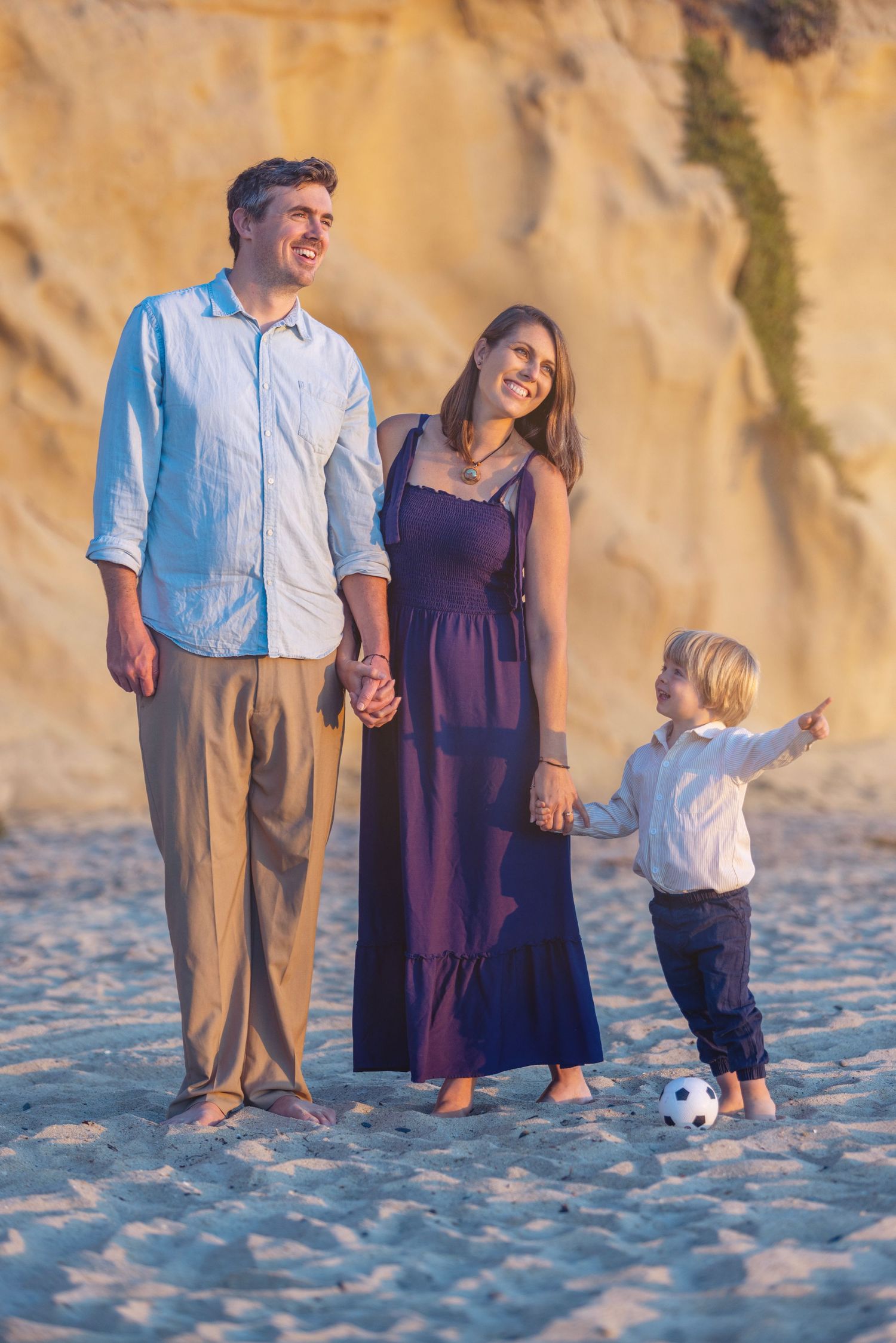Happy family of three enjoying a sunny beach day, parents holding hands with young son playing with soccer ball near sandy cliffs.
