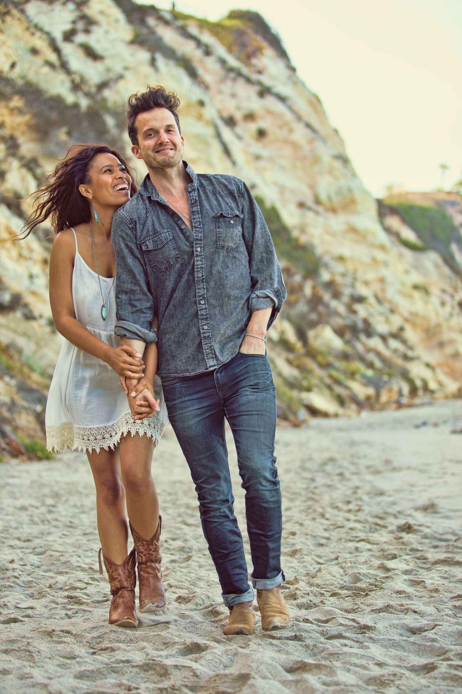 Happy interracial couple walking hand in hand on a sandy beach with rocky cliffs in the background.