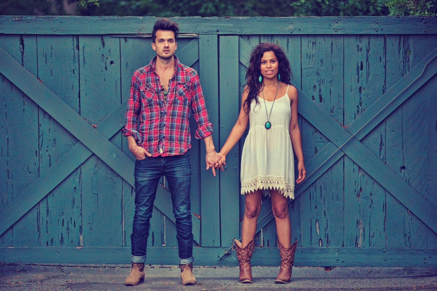 A stylish couple holding hands in front of a rustic teal barn door, wearing bohemian casual outfits.