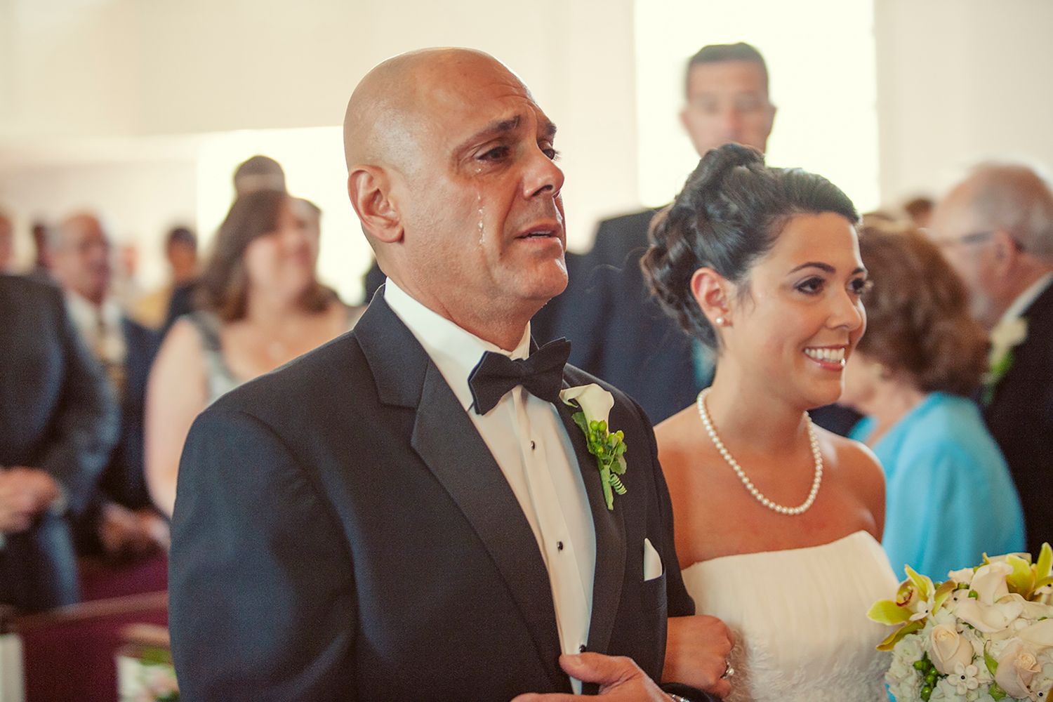Father walks bride down the aisle at wedding ceremony, both smiling with guests in background.