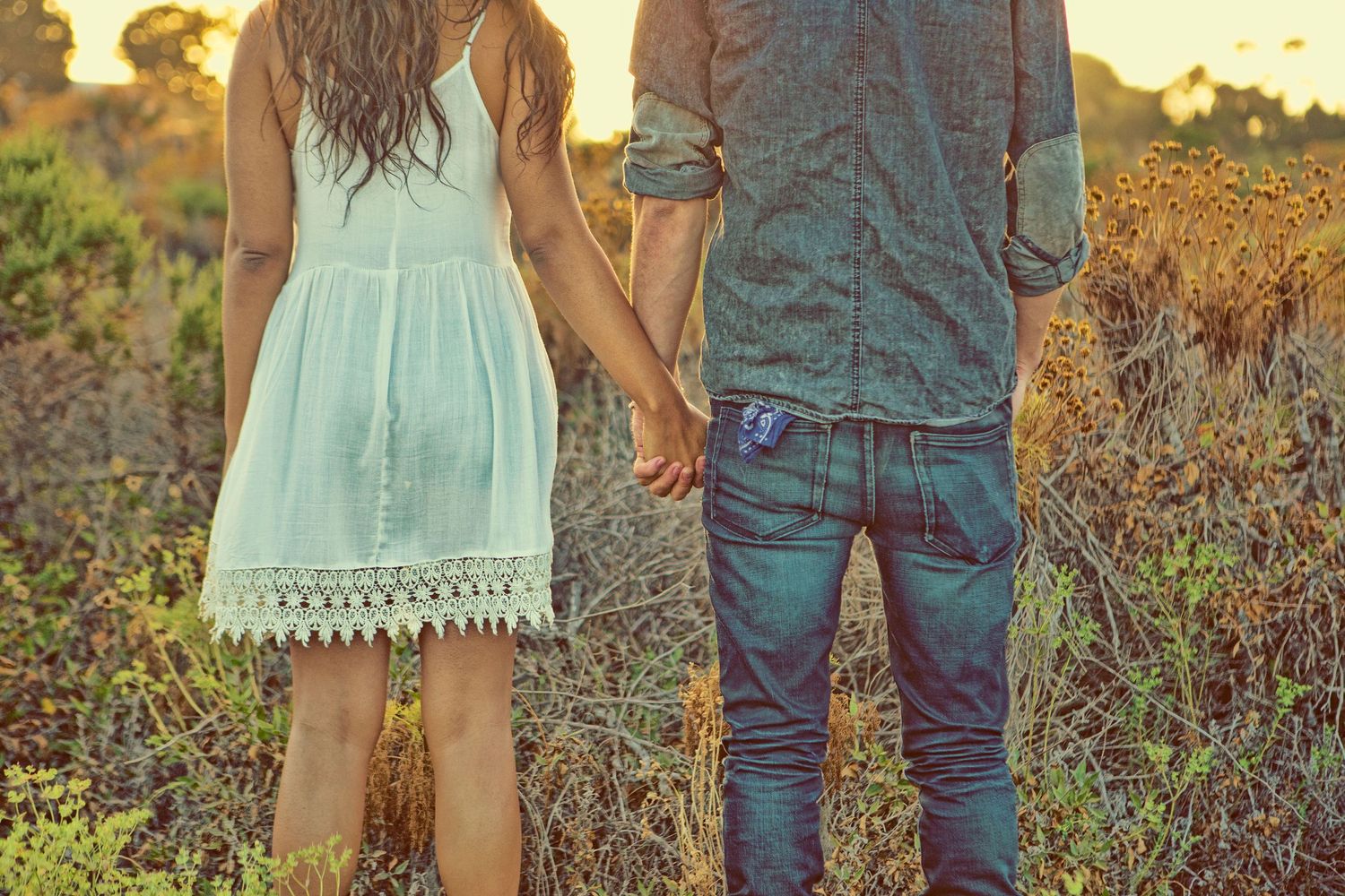 Couple holding hands in a golden field at sunset, woman in white dress and man in jeans.