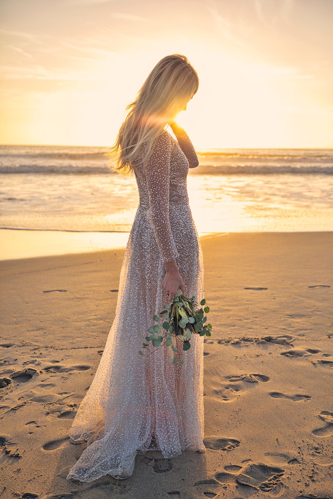 Blonde bride in a flowing lace gown holds a bouquet on a sandy beach at golden sunset.