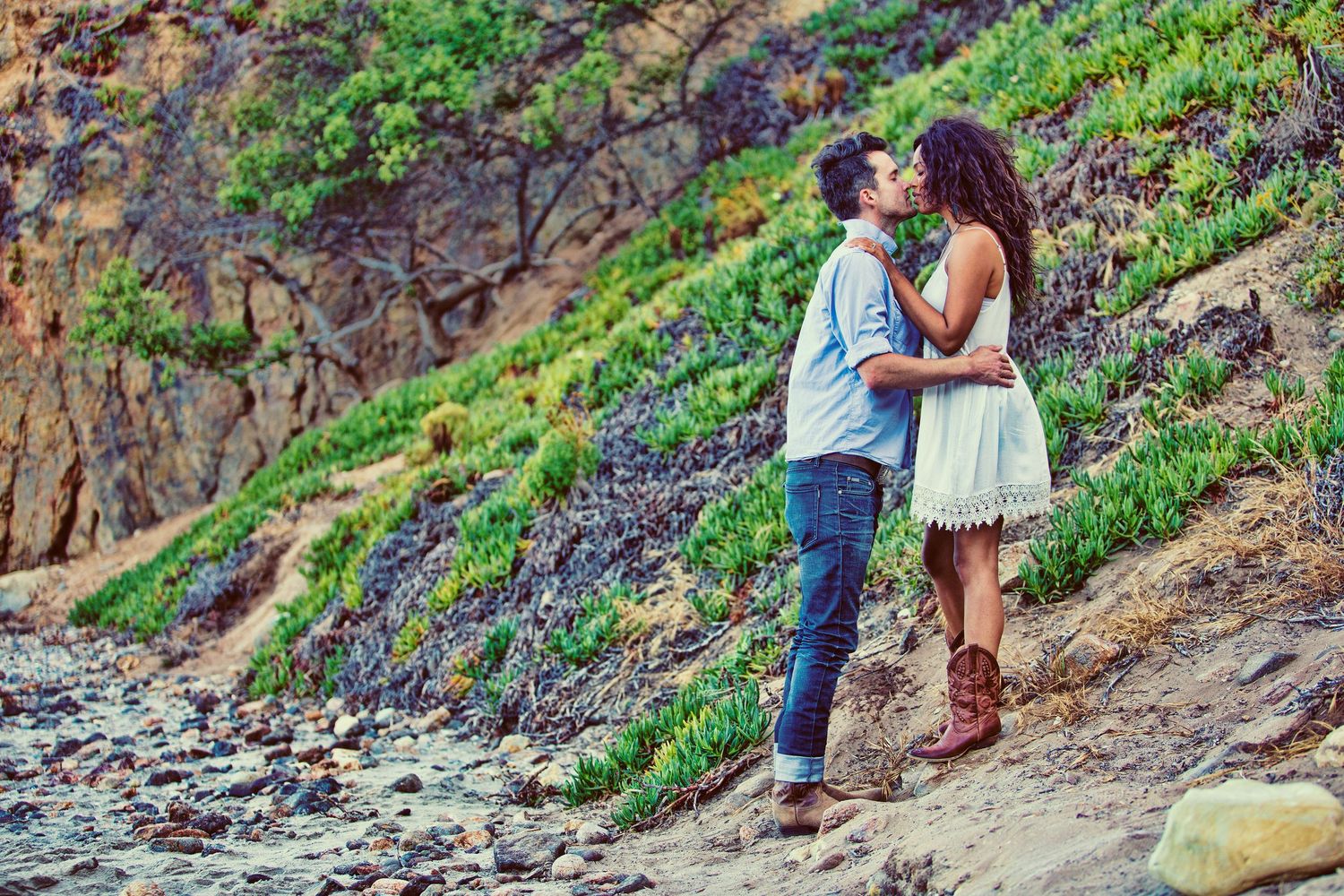 A couple shares a romantic kiss on a rocky beach cliff, woman in white dress and boots, man in blue shirt and jeans.