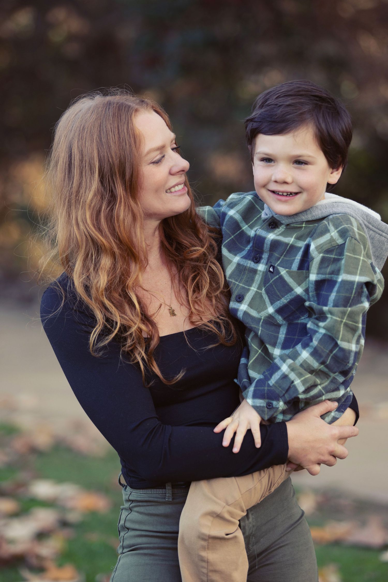 A smiling woman with long red hair holds a happy young boy in a plaid shirt outdoors in autumn.