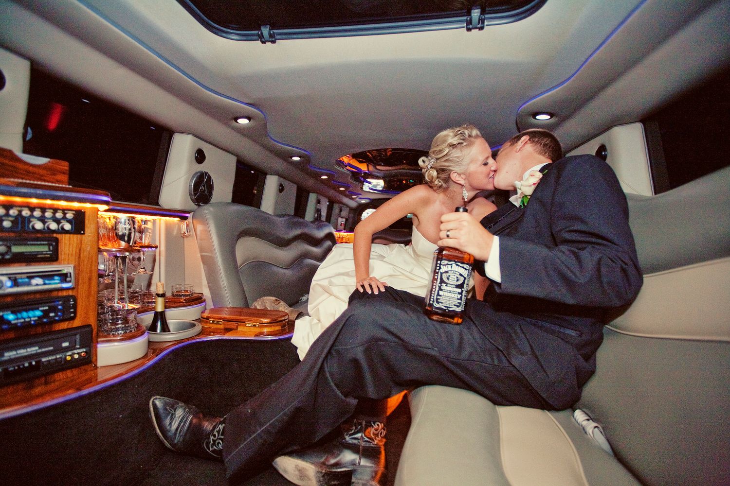Newlywed couple kissing in a luxurious stretch limousine, bride in white dress, groom in suit, holding whiskey bottle.