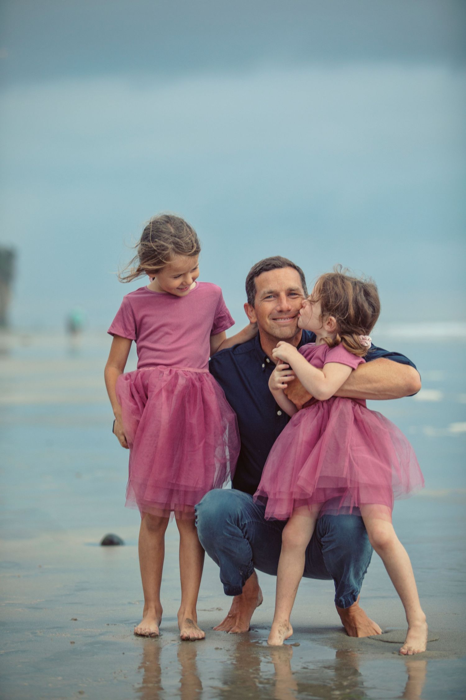 Father crouching on beach with two daughters in matching pink tutu skirts, one kissing his cheek.