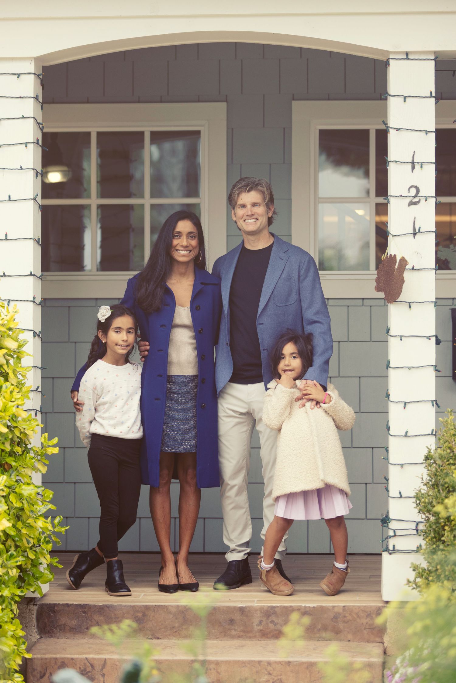 Happy family of four posing on front porch steps of a charming gray house, smiling parents with two young daughters.