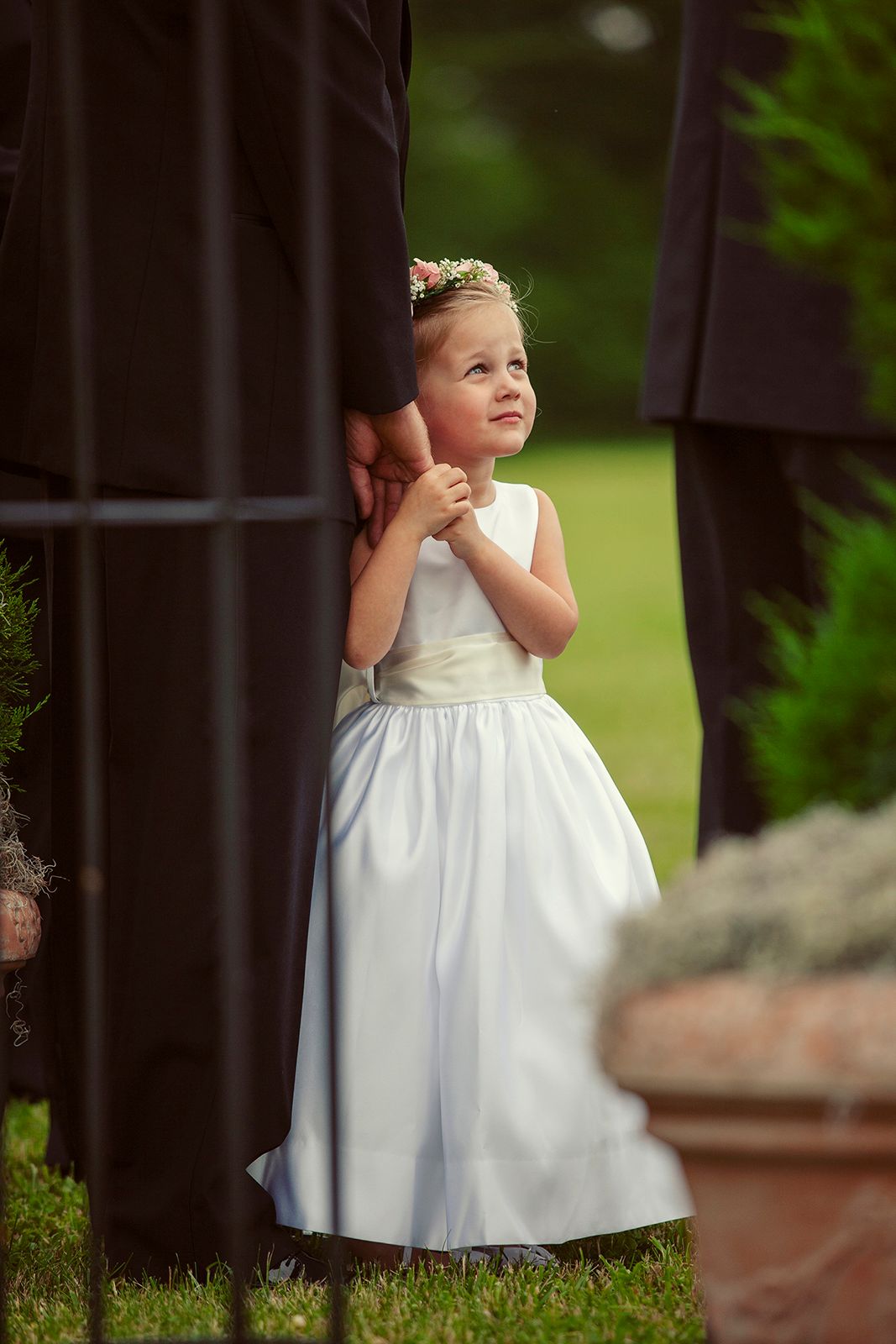 A young flower girl in a white dress and floral crown peeks around a dark pillar during an outdoor wedding ceremony.