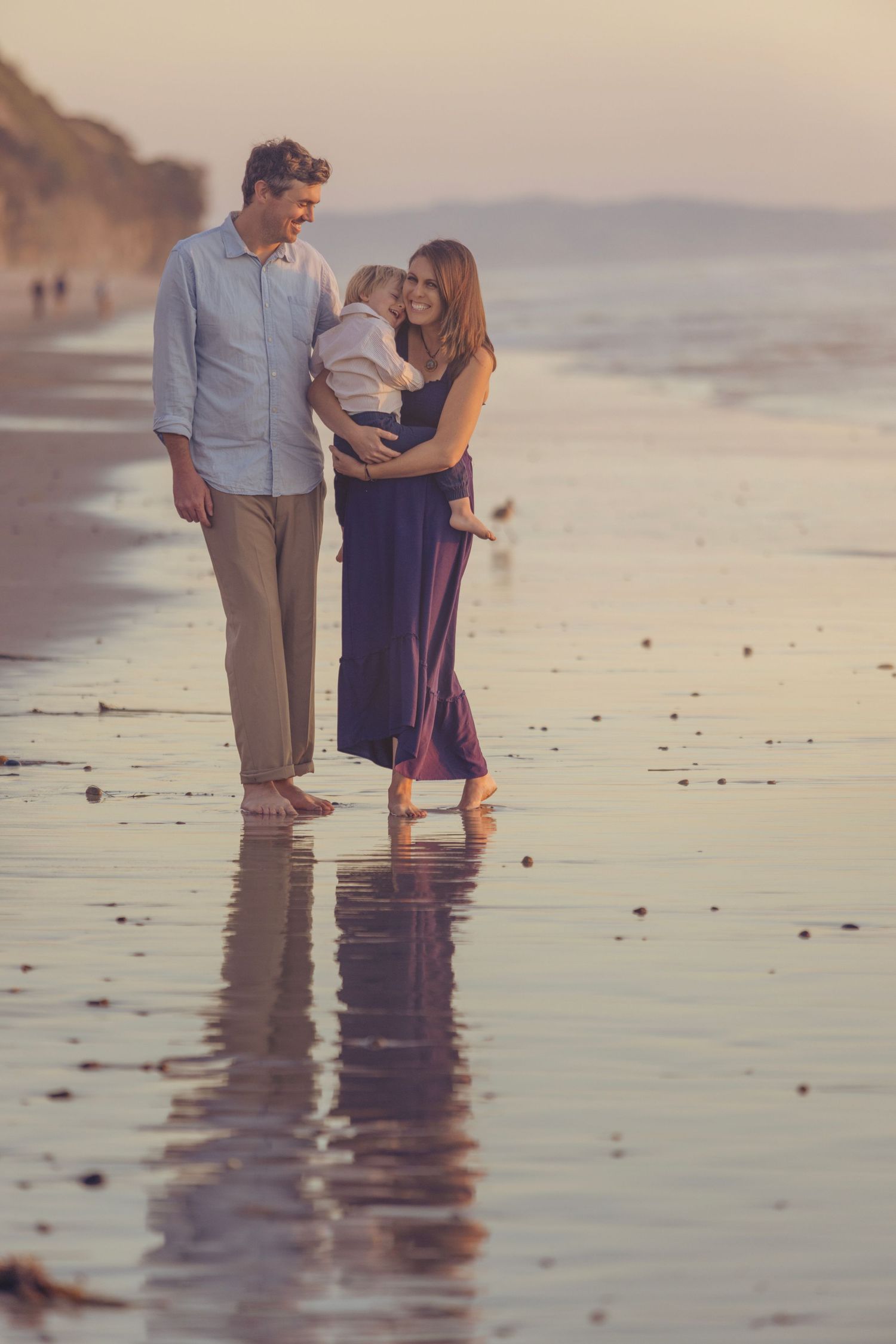 Family of three enjoying a romantic sunset beach walk, with mother holding toddler near ocean waves.