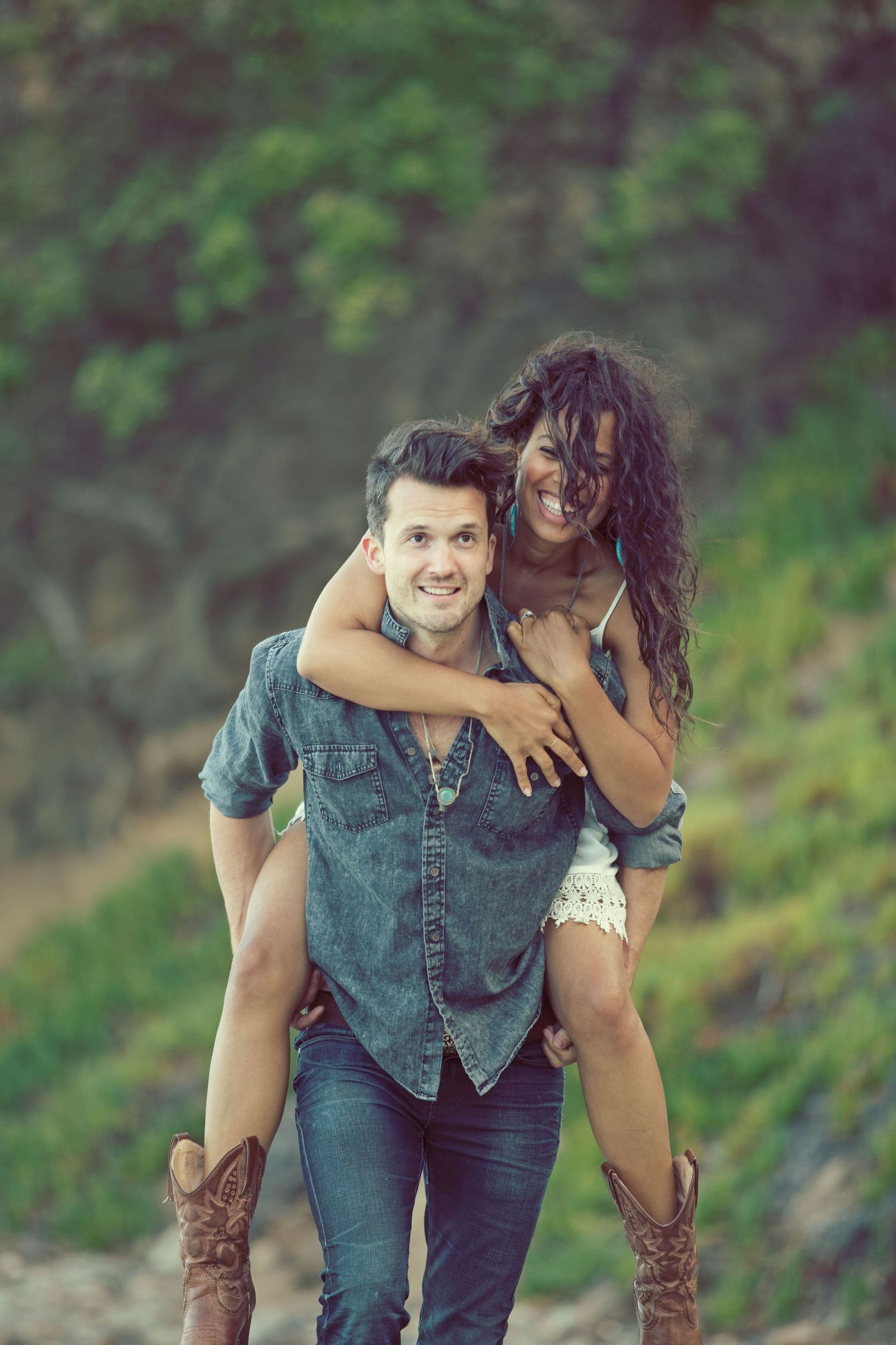 Happy interracial couple piggyback outdoors, man in denim shirt and jeans, woman in white dress smiling.